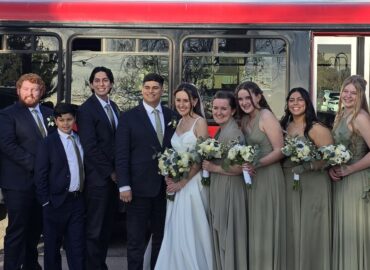 Bride, groom, and wedding party smiling in front of red limo bus