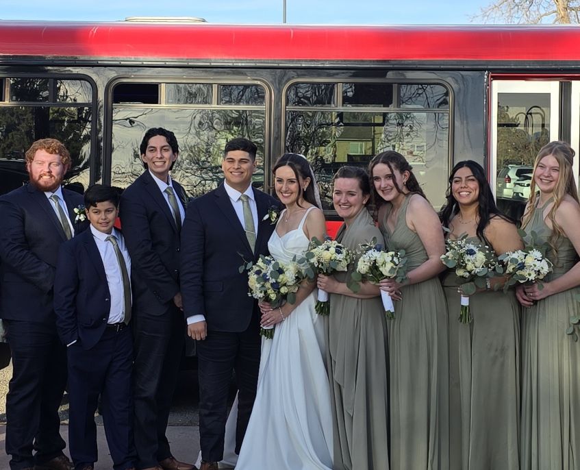 Bride, groom, and wedding party smiling in front of red limo bus