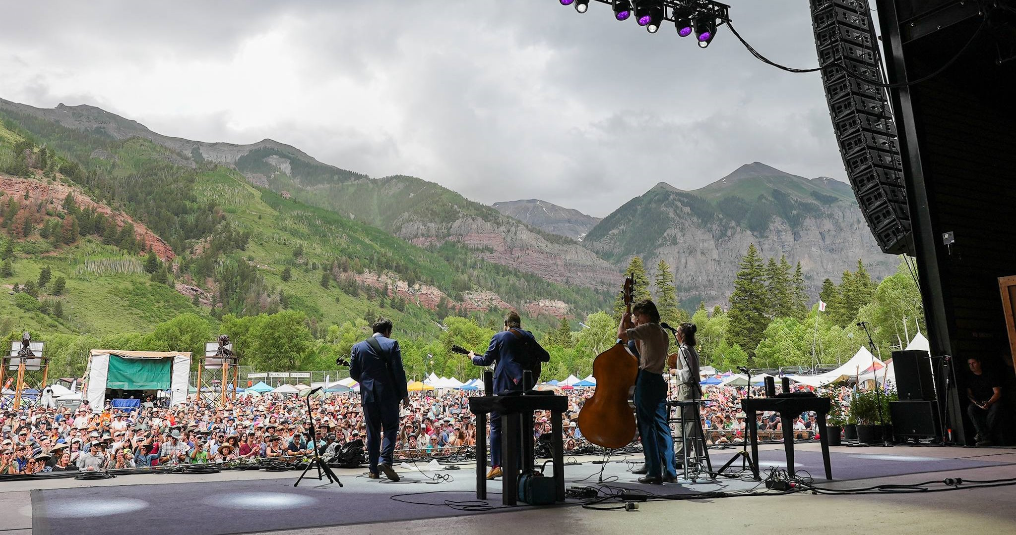 Three musicians on stage in front of a large crowd with green and rocky mountains in the background