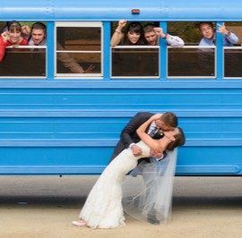 Bride and groom kissing in front of blue bus with people cheering out of the windows