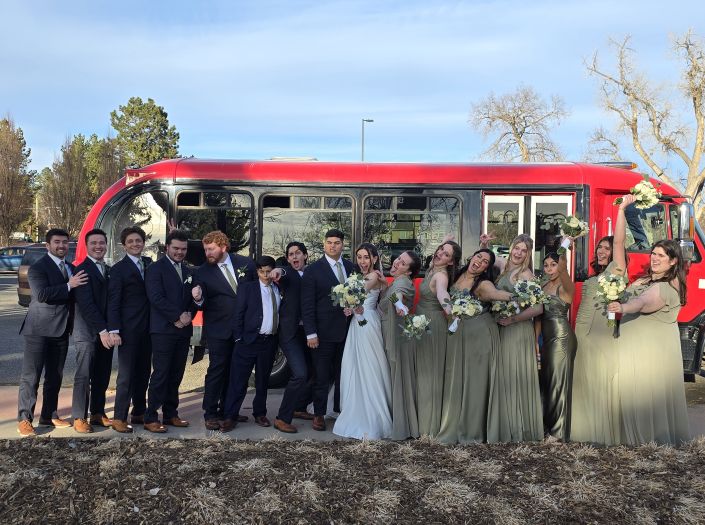 Full wedding party smiling and laughing outside in front of a contoured red limo bus