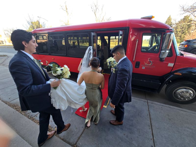 A groomsman and a bridesmaid helping the bride in a white dress board the limo bus, walking on a red carpet